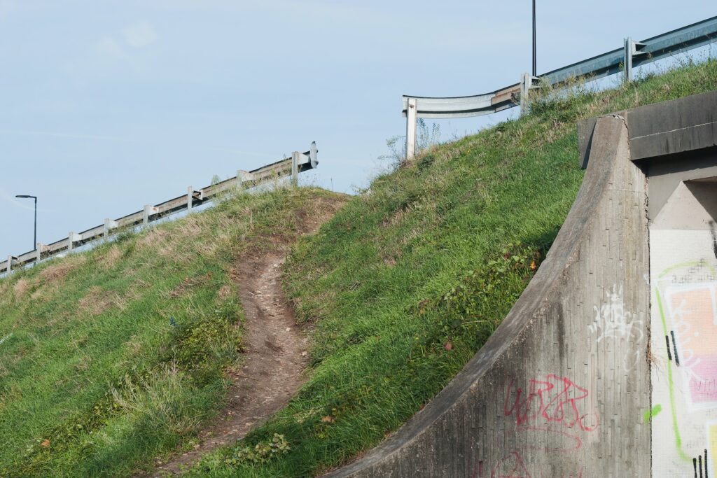 Dirt path leading up a grassy hill and through a hole in a metal guard rail