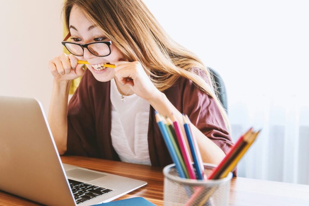 A young woman with glasses is biting a yellow pencil while looking intensely at a laptop screen, with a cup of colorful pencils on her desk, suggesting a moment of stress over the DAM system.